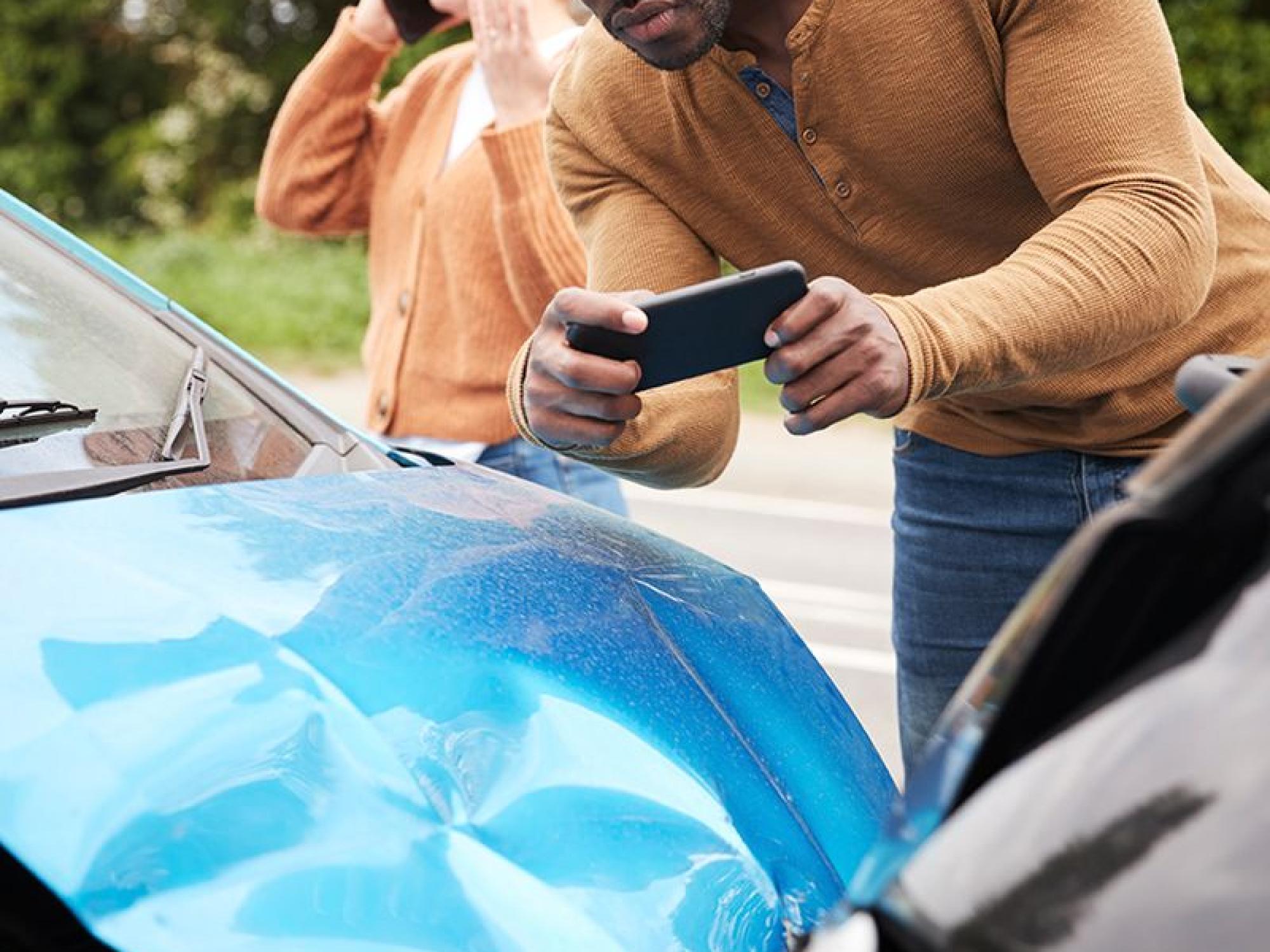 a man standing behind a crashed car, taking a picture with a phone